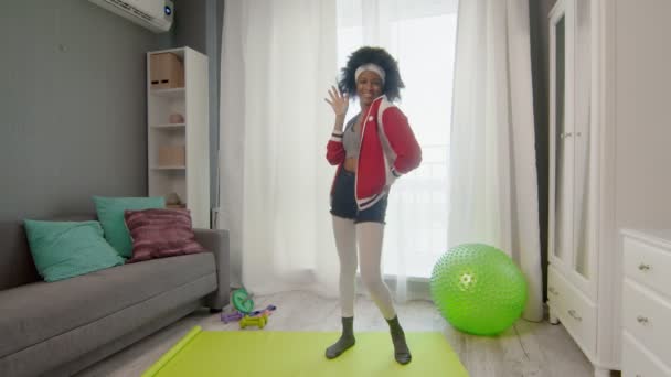 Young african american woman hippy in colourful sportswear with curly afro hairs smiles, looks at the camera and waves her hand — Stock Video