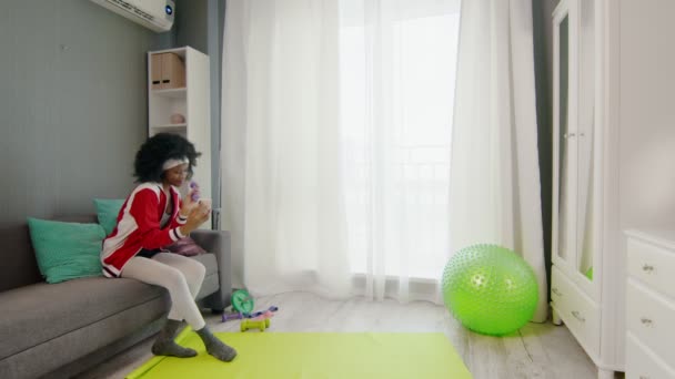 Giovane donna afro americana hippy in abbigliamento sportivo colorato con ricci capelli afro è seduto sul divano, parlando al telefono e mostrando i suoi strumenti sportivi alla fotocamera del telefono — Video Stock