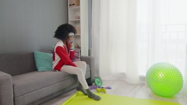 Wanita muda Afrika hippy Amerika dalam pakaian olahraga berwarna-warni dengan rambut afro keriting duduk di sofa, berbicara di telepon dan menunjukkan alat olahraganya di kamera telepon — Stok Video