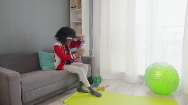 Wanita muda Afrika hippy Amerika dalam pakaian olahraga berwarna-warni dengan rambut afro keriting duduk di sofa, berbicara di telepon dan menunjukkan alat olahraganya di kamera telepon — Stok Video