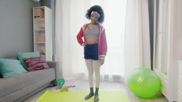 Fiatal vicces afro-amerikai nő hippi színes sportruházat göndör afro haj néz a kamerába, és nem féreg fel gyakorlat a láb — Stock videók