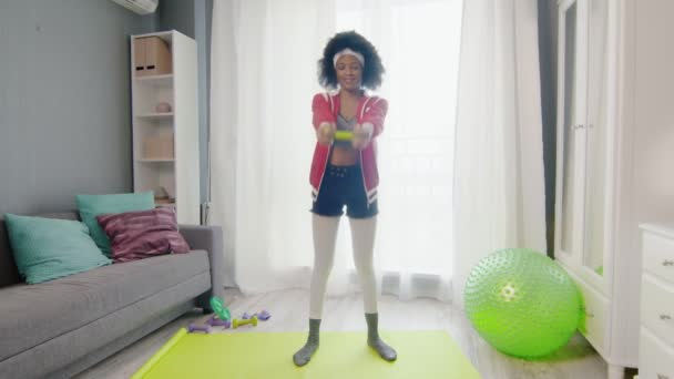 Young funny african american woman hippy in colourful sportswear with curly afro hairs looks at the camera and lifts green dumbel — Stock Video