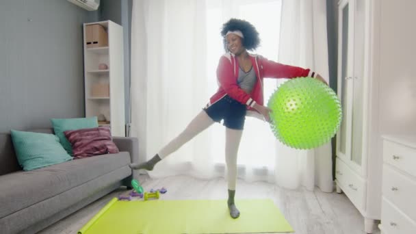 Fiatal vicces afro-amerikai nő hippi színes sportruházat göndör afro szőrszálak néz a kamerába, tartja fitness labda a kezében, és felemeli a lábát — Stock videók