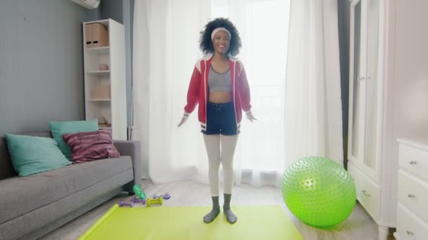 Nuori onnellinen afrikkalainen amerikkalainen nainen hippi värikkäissä urheiluvaatteissa, joissa on kiharat afrokarvat, katsoo kameraan ja lämmittää liikuntaa. — kuvapankkivideo