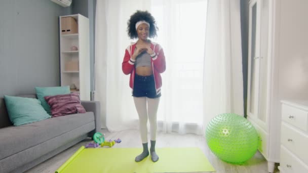 Young african american woman hippy in colourful sportswear with curly afro hairs looks at the camera and does squats exercise — Stock Video