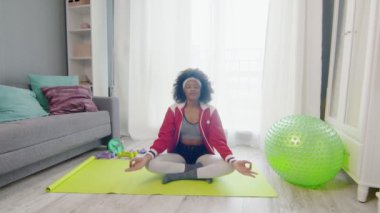Afro-Amerikan kadın hippi renkli spor kıyafetleri ve kıvırcık afro saçlarıyla lotus pozu ve meditasyonlarda oturuyor.