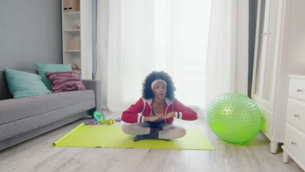 Young african american woman hippy in colourful sportswear with curly afro hairs sits in lotus pose, looks at the camera and does stretch exercise — Stock Video