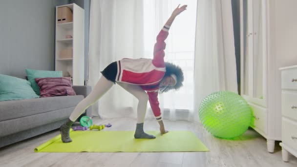 Jeune femme afro-américaine hippie en vêtements de sport colorés avec des cheveux afro bouclés pose de yoga triangle — Video