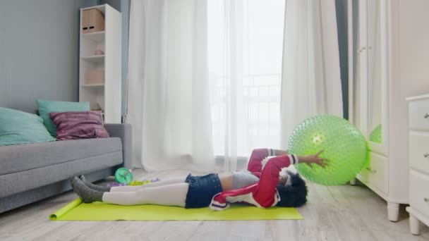 Young african american woman hippy in colourful sportswear with curly afro hairs lays on mat and does exercise with fitness ball at home — Stock Video