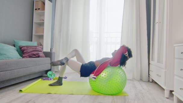 Young african american woman hippy in colourful sportswear with curly afro hairs lays on fitness ball and does abs exercises crunches at home — Stock Video