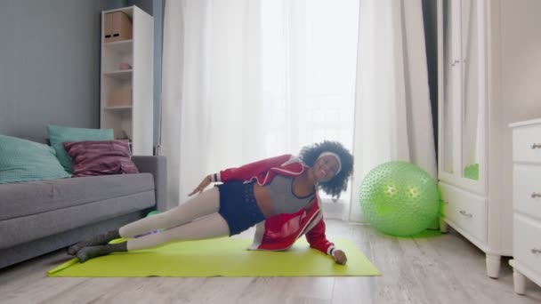 Young african american woman hippy in colourful sportswear with curly afro hairs looks at the camera, stays in side plank and lifts hand — Stock Video
