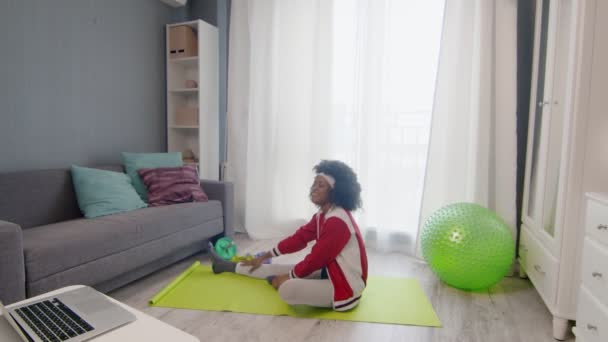 Joven mujer afroamericana hippy en ropa deportiva colorida con pelos afro rizados hace ejercicio de estiramiento y mira video tutorial de fitness en línea en el ordenador portátil — Vídeos de Stock