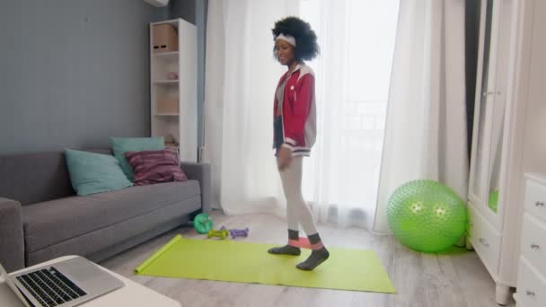 Wanita muda afrika hippy dengan pakaian olahraga berwarna-warni dengan rambut afro keriting melakukan latihan dengan kaki dengan pita elastis dan jam tangan video kebugaran tutorial online pada komputer laptop — Stok Video