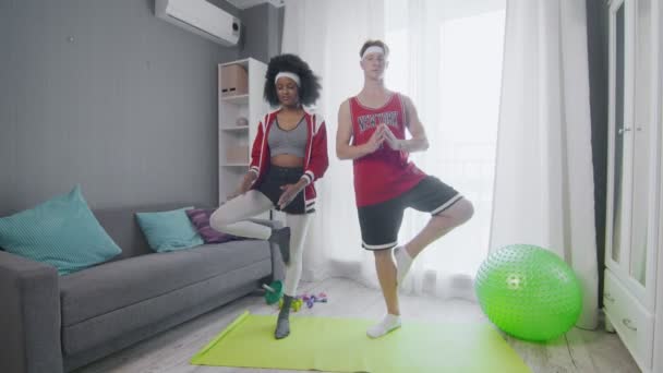 Ridiculous Caucasian man and african american woman newcomers in sport in colourful sportswear does tree pose — Stock Video