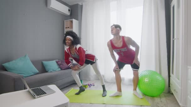 Zabawny styl retro para kaukaski mężczyzna i afro kobieta robi ćwiczenia i ogląda fitness wideo samouczek online na laptopie — Wideo stockowe