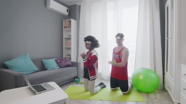 Engraçado casal estilo retro de homem caucasiano e mulher afro fica de joelhos e levanta halteres enquanto assiste vídeo tutorial fitness on-line no computador portátil — Vídeo de Stock