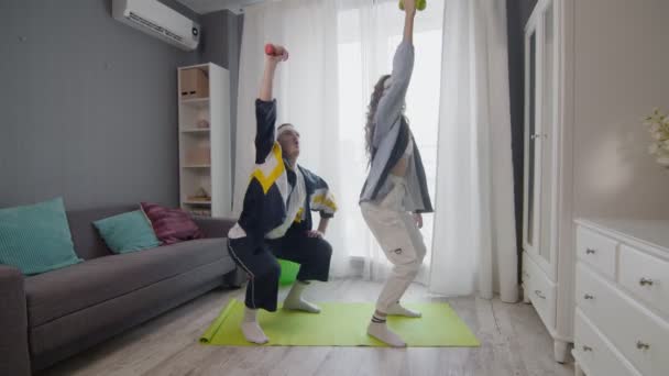 Pasangan muda bahagia yang sehat pria dan wanita bekerja di rumah. Mereka melakukan squat dan mengangkat dombbells up — Stok Video