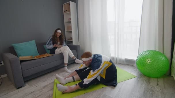 Wanita muda bergaya retro makan pizza dan berbicara di telepon sementara pacarnya melakukan latihan peregangan di rumah — Stok Video