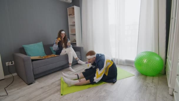 Wanita muda bergaya retro makan pizza dan menggunakan telepon sementara pacarnya melakukan latihan peregangan di rumah — Stok Video