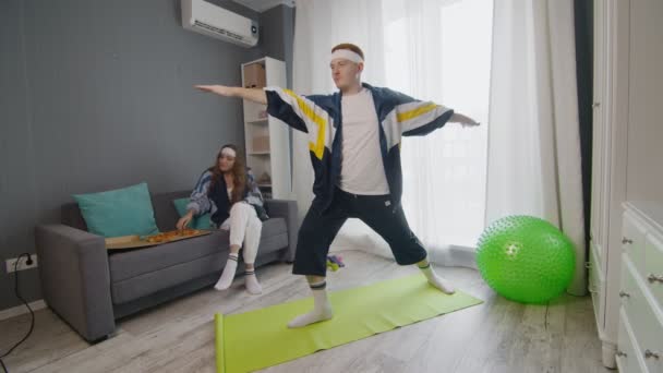 Retro snygg ung kvinna äter pizza medan hennes pojkvän gör yoga krigare poserar hemma — Stockvideo