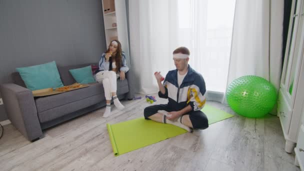 Ретро стильная молодая женщина ест пиццу, в то время как ее парень сидит на коврике и делает упражнения с гантели дома — стоковое видео