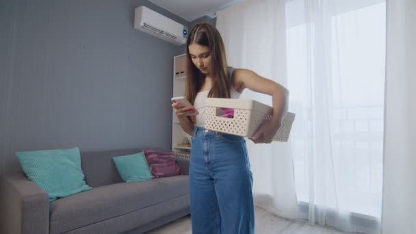 Веселая молодая женщина держит корзину одежды и с помощью телефона — стоковое видео
