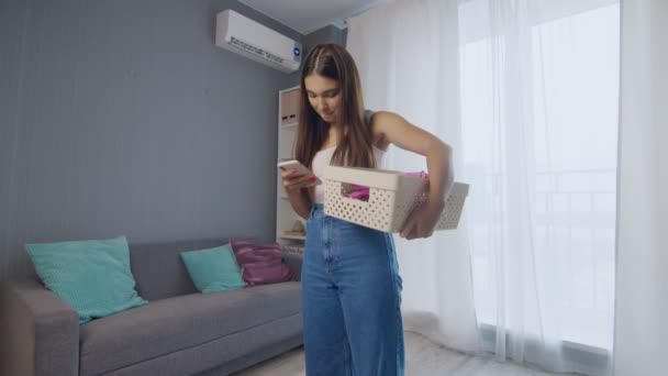 Веселая молодая женщина держит корзину с одеждой, пользуется телефоном и смотрит в камеру — стоковое видео