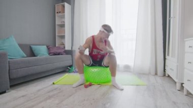 Retro stil bir genç adam bir fitness topunun üzerine oturur ve evde telefonuyla müzik dinler.
