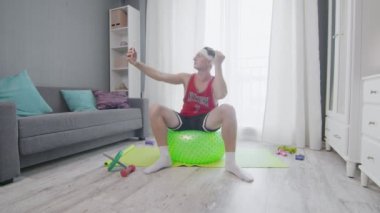 Retro stil genç adam spor yapmadan önce bir fitness topuna oturur ve selfie çeker.