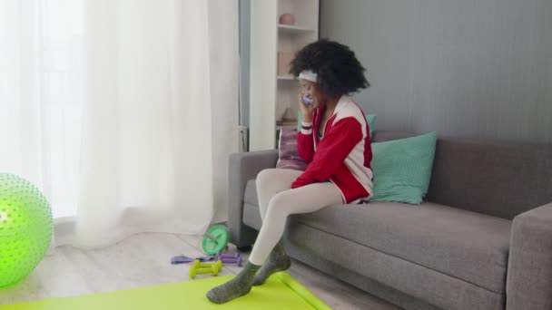 Young african american woman hippy in colourful sportswear with curly afro hairs is sitting on couch, talking on the phone and showing her sport tools at the phone camera — Stock Video
