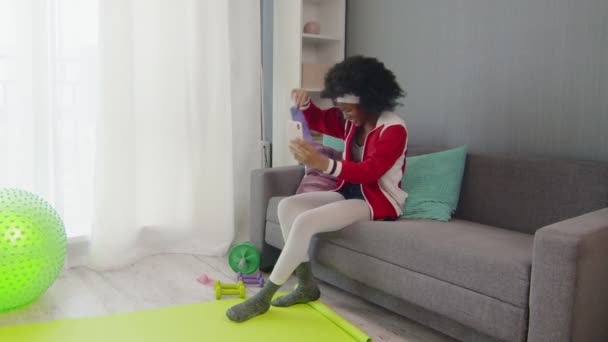 Молодая африканская американка хиппи в разноцветной спортивной одежде с кудрявыми афро-волосами сидит на диване, разговаривает по телефону и показывает свои спортивные инструменты на камеру телефона — стоковое видео