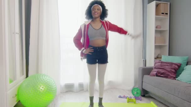 Nuori hauska afrikkalainen amerikkalainen nainen hippi värikkäissä urheiluvaatteissa, joissa on kiharat afrokarvat, katsoo kameraan ja tekee venytysharjoituksia. — kuvapankkivideo