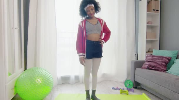 Muda lucu african Amerika perempuan hippy dalam warna-warni sportswear dengan rambut afro keriting menatap kamera dan melakukan worm up latihan untuk kaki — Stok Video