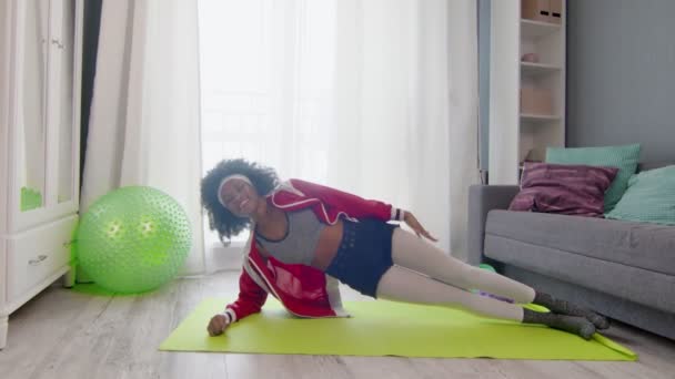 Young african american woman hippy in colourful sportswear with curly afro hairs looks at the camera, stays in side plank and lifts hand — Stock Video