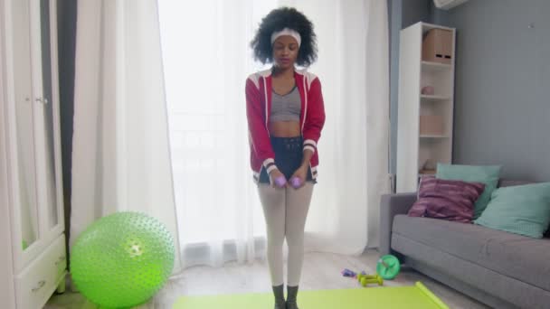 Young african american woman hippy in colourful sportswear with curly afro hairs looks ant the camera and lifts dumbbells at home — Stock Video