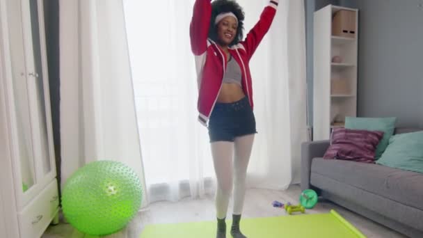 Young african american woman hippy in colourful sportswear with curly afro hairs looks ant the camera and lifts dumbbells up at home. — Stock Video