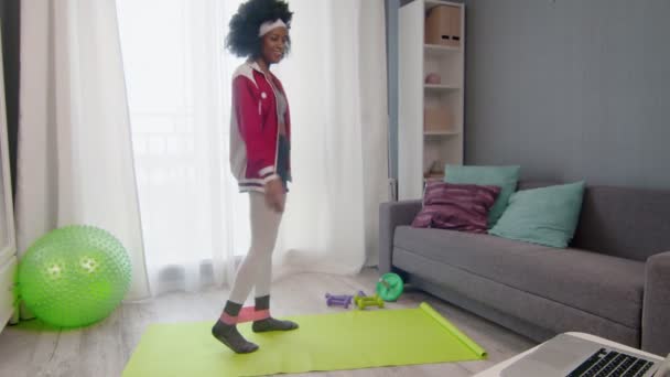 Nuori afrikkalainen amerikkalainen nainen hippi värikkäissä urheiluvaatteissa, joissa on kiharat afrokarvat, käyttää jalkoja joustavalla bändillä ja katsoo fitness-videon opetusohjelmaa verkossa kannettavalla tietokoneella — kuvapankkivideo