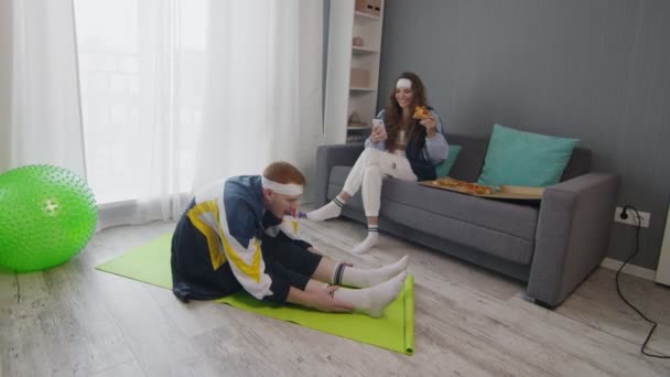 Retro snygg ung kvinna äter pizza och använder telefon medan hennes pojkvän gör stretching motion hemma — Stockvideo