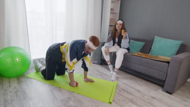 Retro elegante joven mujer comiendo pizza mientras su novio haciendo ejercicio con la mancuerna en casa — Vídeo de stock