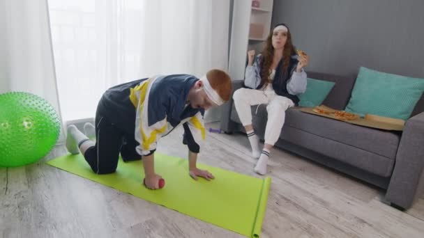 Zoom out retro stylish young woman eating pizza while her boyfriend doing exercise with dumbbell at home — Stock Video