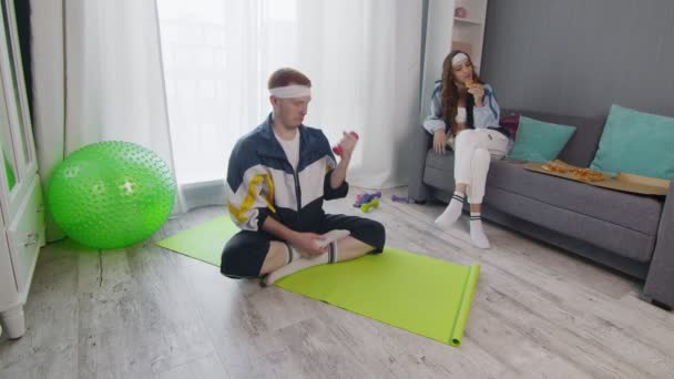 Retro stylish young woman eating pizza while her boyfriend sitting on mat and doing exercise with dumbbell at home — Stock Video