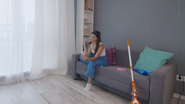 Уставшая молодая женщина после уборки дома одна сидит на тренере и пользуется телефоном — стоковое видео