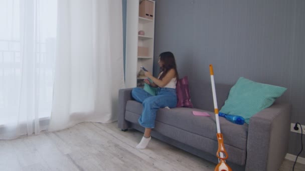 Moe jonge vrouw na het schoonmaken van het huis alleen zit op de coach en maakt gebruik van telefoon — Stockvideo