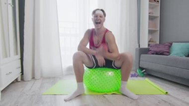 Komik bir genç adam spor topuyla antrenman yapıyor ve evinde dambılı kaldırıyor.