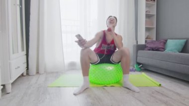 Retro stil genç adam bir fitness topunun üzerine oturur, telefonuyla müzik dinler ve mikrofondaki gibi dambılda şarkı söyler.