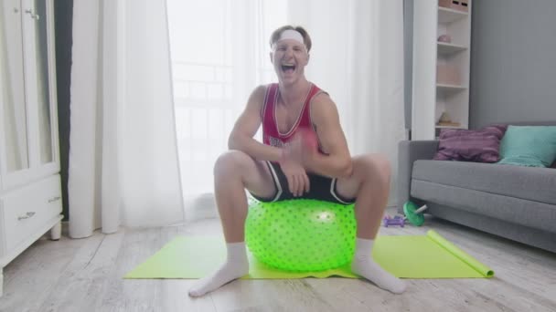 Vicces fiatalember edz egy fitnesz labdán és súlyzót emel a házában. — Stock videók