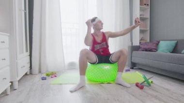 Retro stil genç adam spor yapmadan önce bir fitness topuna oturur ve selfie çeker.