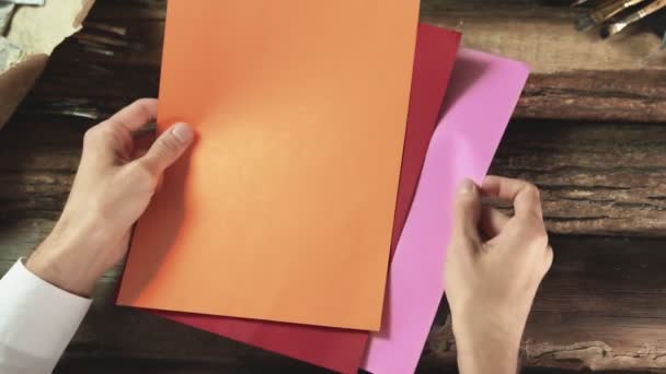 Mężczyzna posiada trzy kolorowe papiery, drewniany stół na tle — Wideo stockowe