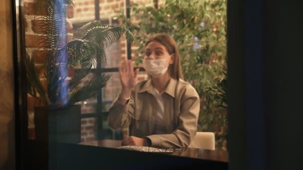 Mujer joven con máscara protectora se sienta en la cafetería cerca de la ventana y las olas de la mano a su amigo fuera — Vídeos de Stock