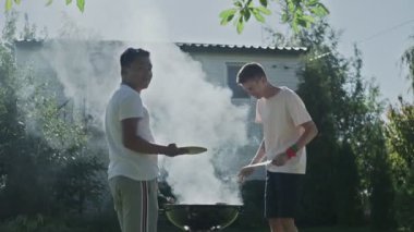 İki mutlu adam ızgarada domuz pastırması ve sosis pişiriyor.